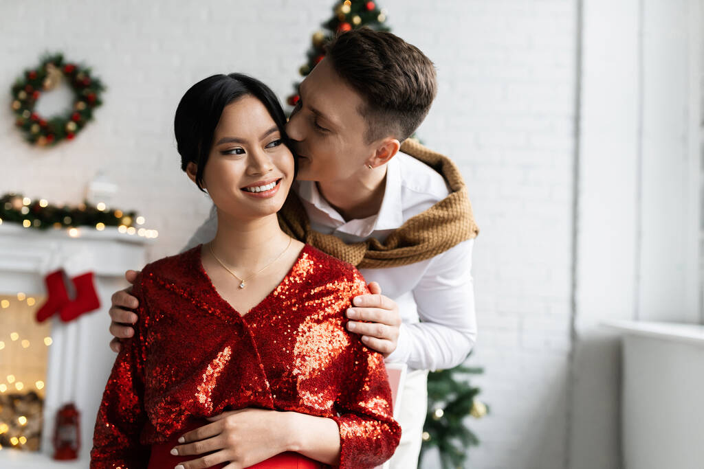 young man kissing pregnant asian woman in red and shiny blouse near Christmas decor on blurred background - Photo, Image