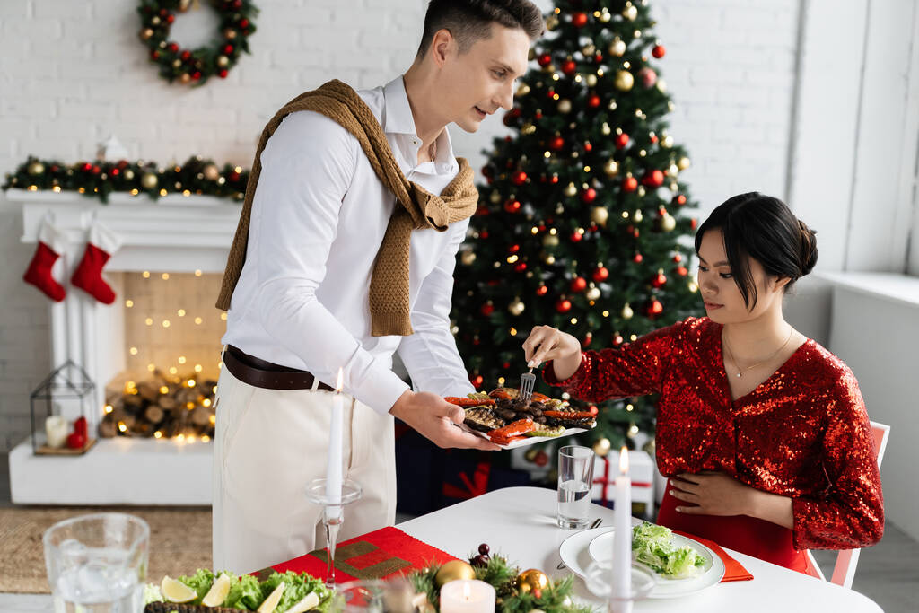 クリスマスツリーの近くでロマンチックな夕食の間に妊娠中のエレガントなアジアの女性の近くにグリル野菜を保持男  - 写真・画像