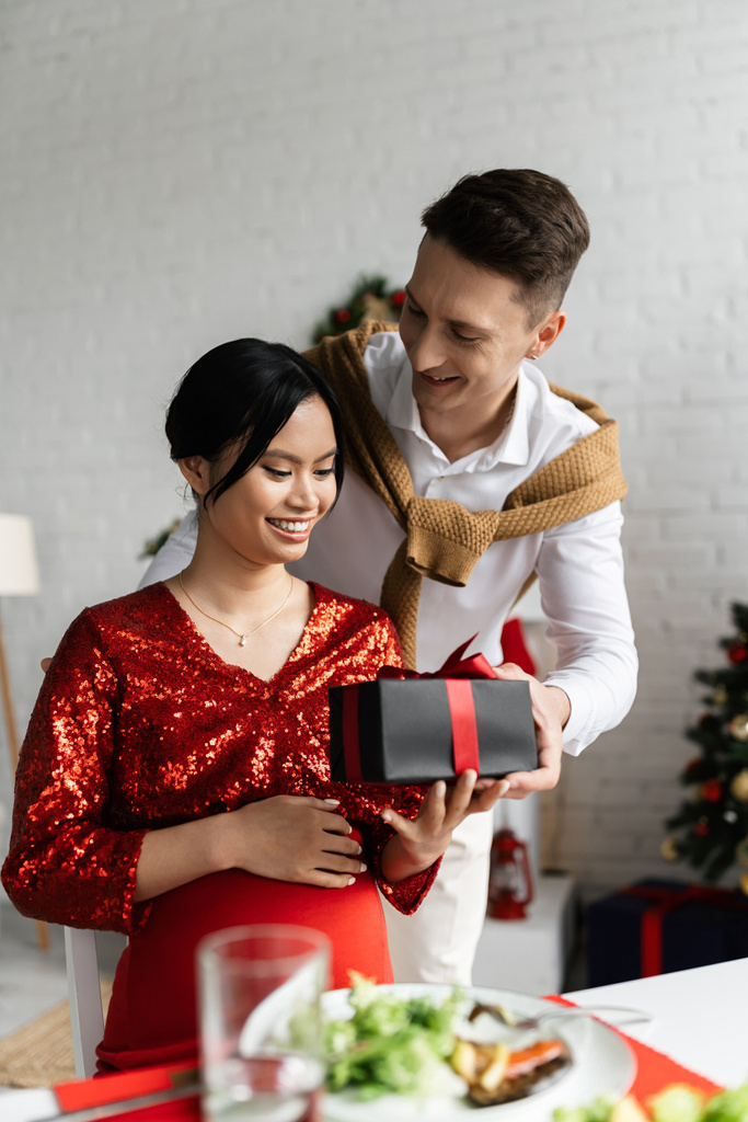 junger Mann beschenkt schwangere und elegante asiatische Ehefrau mit Weihnachtsgeschenk - Foto, Bild