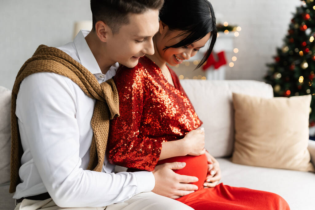 молодий чоловік обіймає животик вагітної і щасливої азіатки під час святкування Різдва вдома
 - Фото, зображення