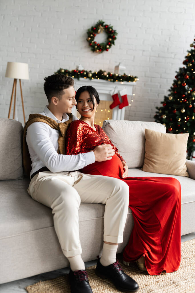 joven tocando vientre de embarazada asiático esposa en festivo ropa en acogedor sofá cerca borrosa Navidad decoración - Foto, Imagen