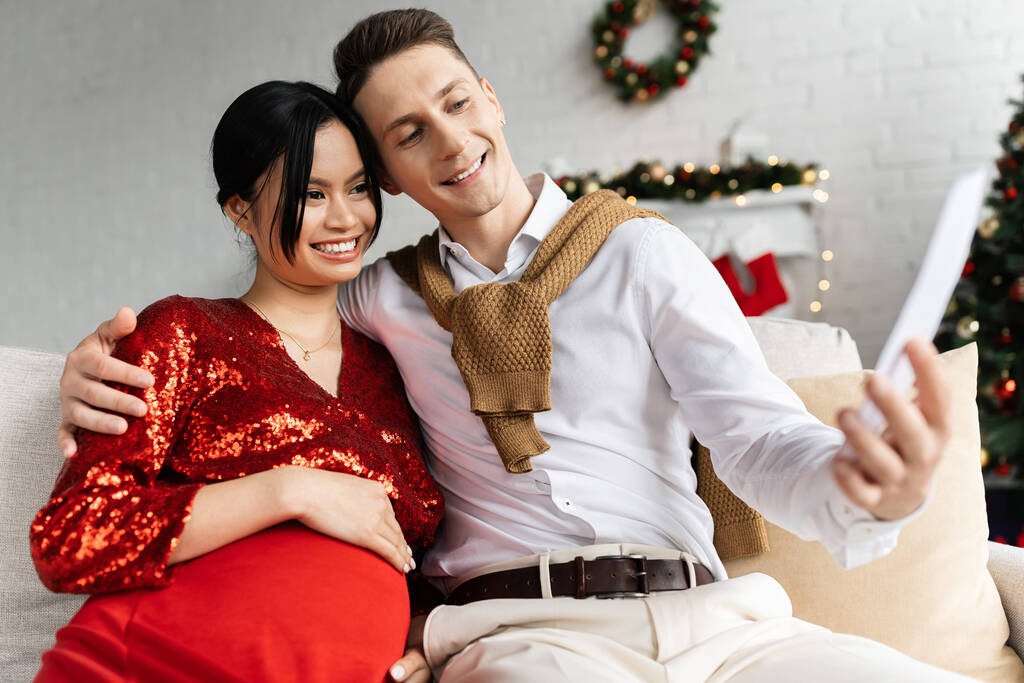 raskaana ja tyylikäs aasialainen nainen hymyilevä aviomies etsii hämärtynyt kortti kotona joulun sisustus - Valokuva, kuva