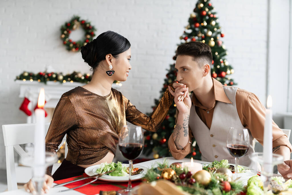 tetovaný muž líbání ruku asijské manželky v elegantním oblečení při romantické večeři na Vánoce doma - Fotografie, Obrázek