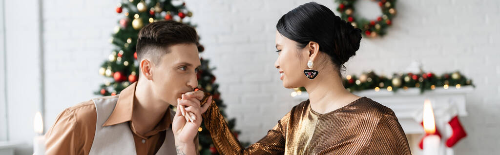 若い男がクリスマスのお祝いの間にリビングルームでエレガントなアジアの妻の手にキス,バナー - 写真・画像