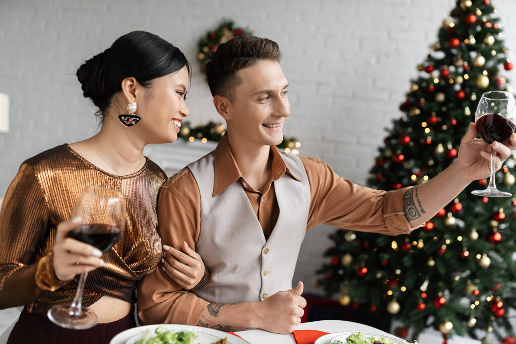 glimlachende man toasten met wijnglas terwijl vieren Kerstmis met gelukkig aziatische vrouw  - Foto, afbeelding