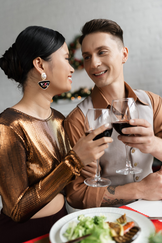 radosna wieloetniczna para w eleganckim stroju patrząca na siebie i brzęcząca kieliszkami wina podczas romantycznej kolacji - Zdjęcie, obraz