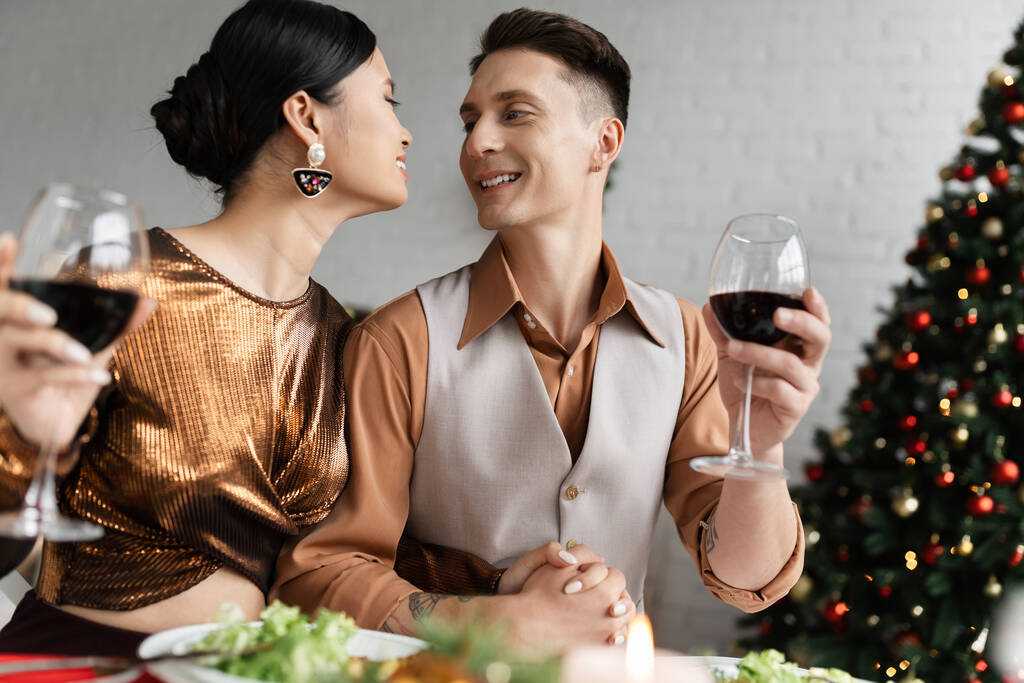 międzyrasowa para w świątecznych ubraniach trzymająca się za ręce i uśmiechająca się do siebie trzymając kieliszki do wina - Zdjęcie, obraz