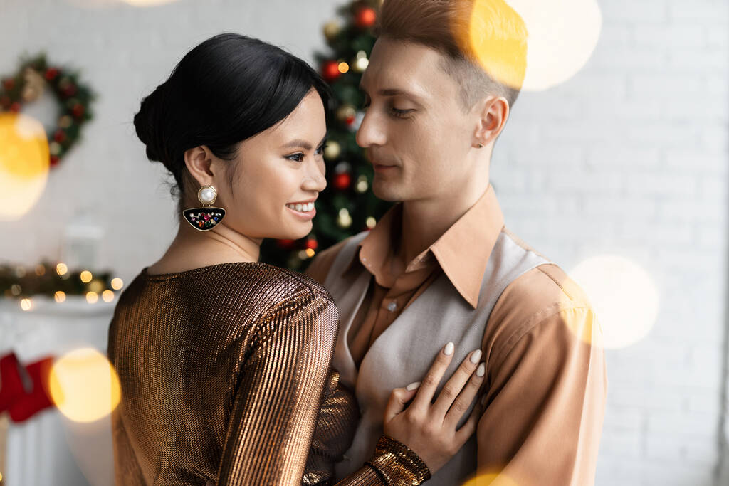 Esmer, Asyalı, şenlikli elbiseli bir kadın. Evdeki Noel kutlaması sırasında genç kocanın yanında gülümsüyor. - Fotoğraf, Görsel