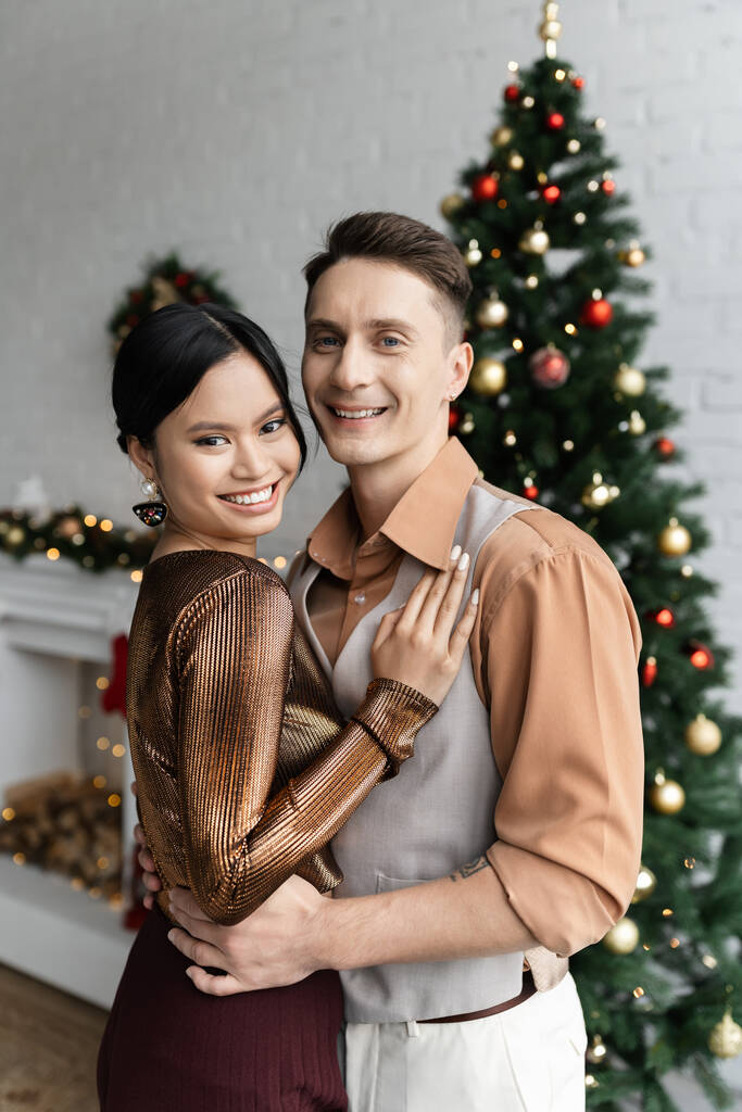 cheerful asian woman and man in festive outfits hugging near Christmas tree  - Photo, Image