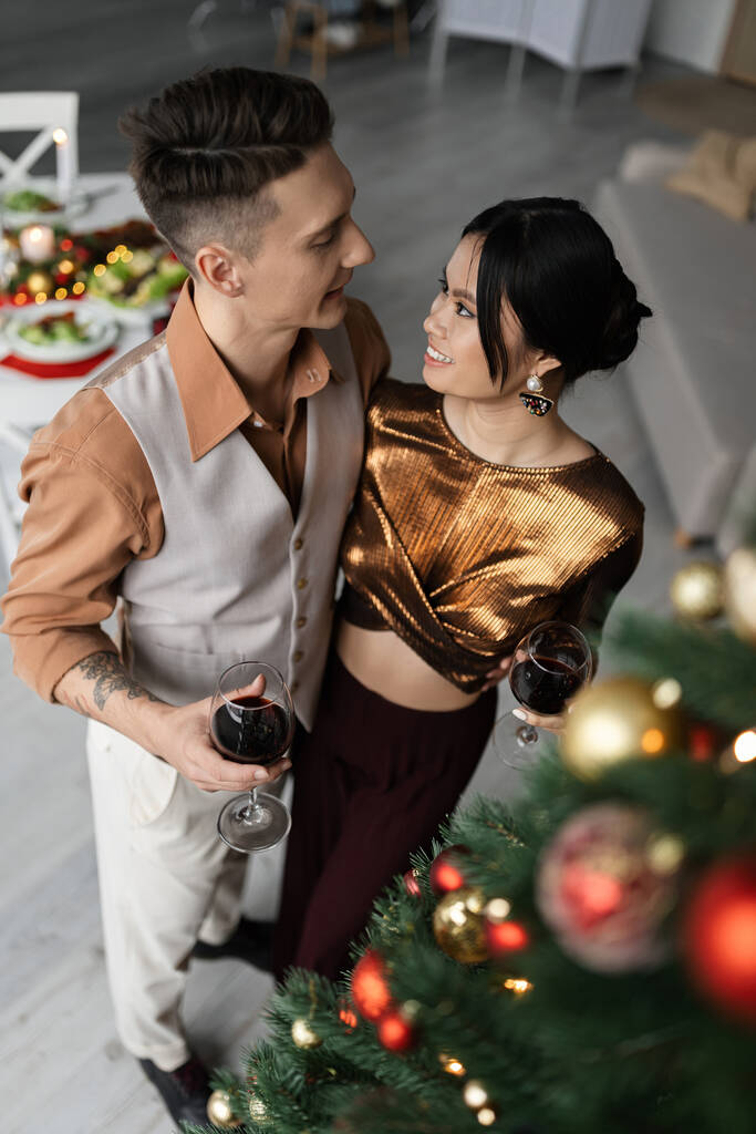 wysoki kąt widzenia szczęśliwy międzyrasowy para w świąteczne stroje trzymając kieliszki wina w pobliżu urządzone choinki  - Zdjęcie, obraz