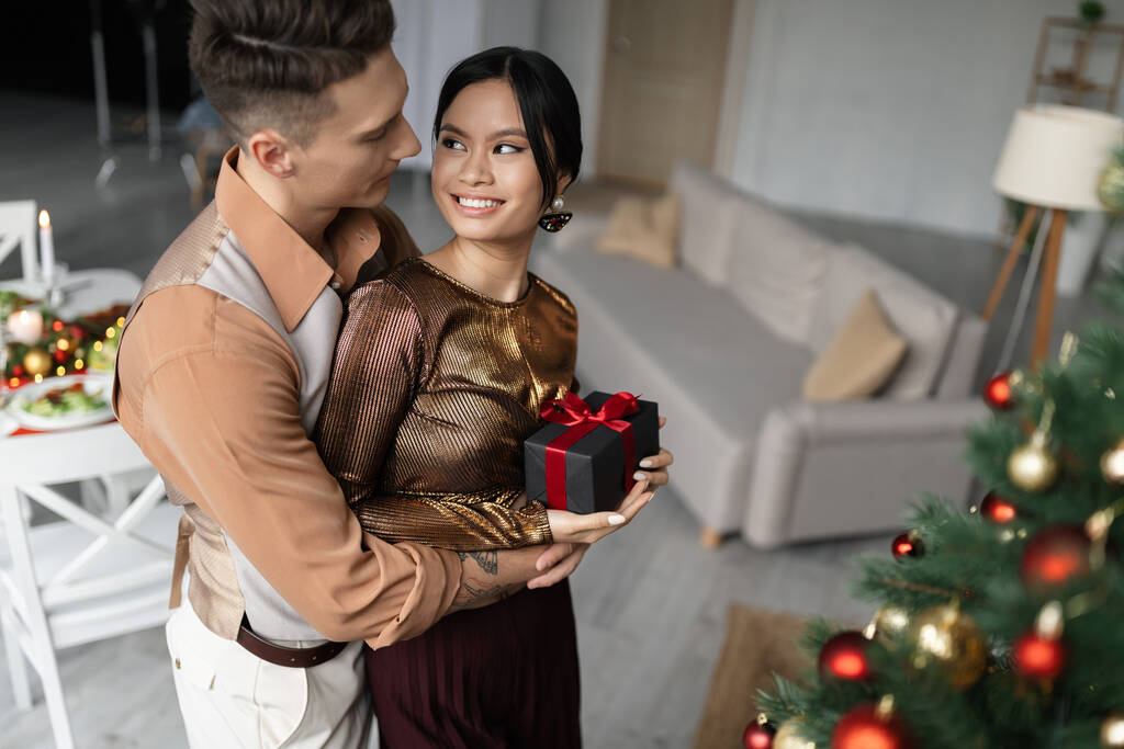 счастливый и татуированный мужчина обнимает азиатскую жену, держа рождественский подарок  - Фото, изображение