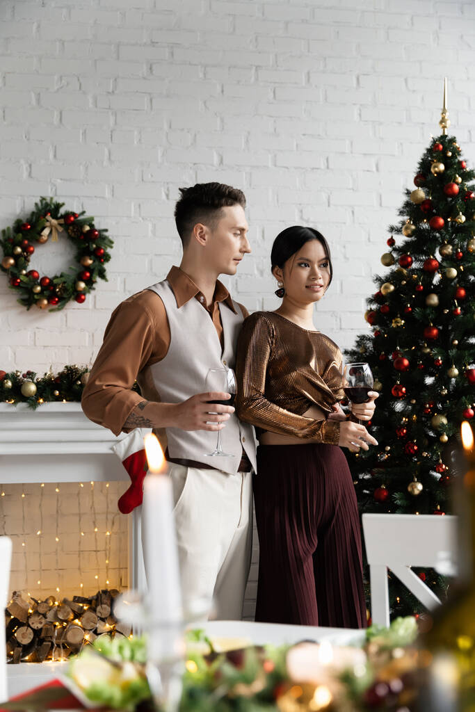 πολυεθνική σύζυγος και σύζυγος κρατώντας ποτήρια κρασιού κατά τη διάρκεια της γιορτής των Χριστουγέννων  - Φωτογραφία, εικόνα