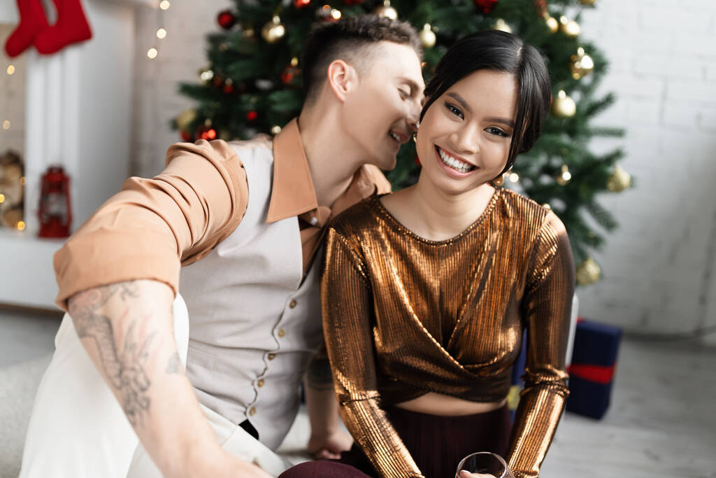 alegre homem sussurrando no ouvido de feliz asiático esposa enquanto sentado sob Natal árvore  - Foto, Imagem