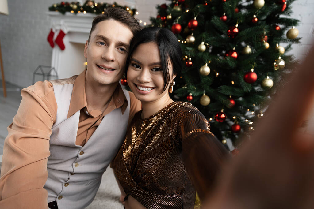 pareja interracial sonriendo y mirando a la cámara mientras se sienta bajo el árbol de Navidad  - Foto, imagen