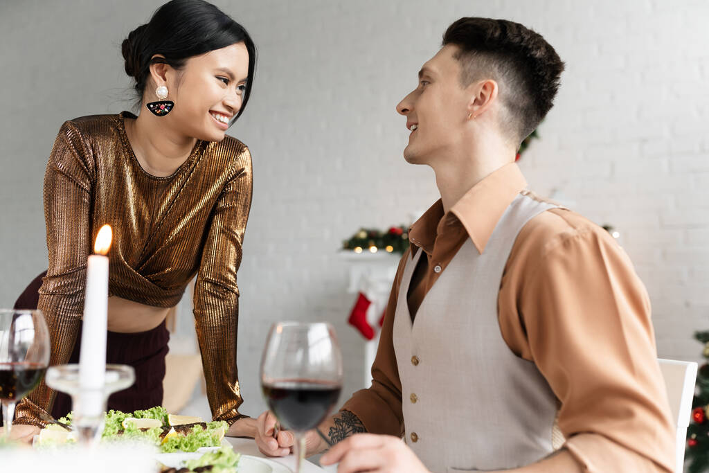 vrolijk interraciaal paar kijken naar elkaar in de buurt feestelijke maaltijd en glazen wijn tijdens Kerstmis  - Foto, afbeelding
