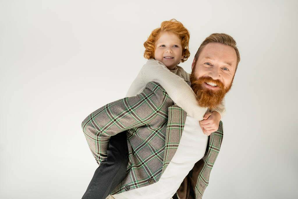Улыбающийся рыжеволосый ребенок, цепляющийся за бородатого папу, изолированного от серого  - Фото, изображение
