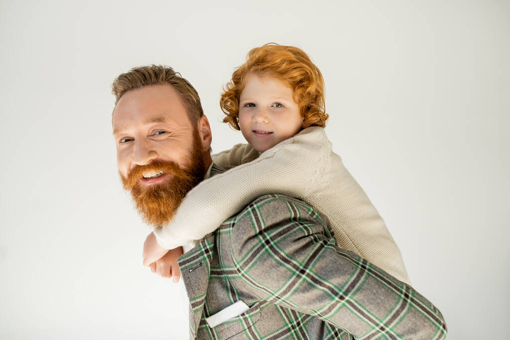 Lächelnder rothaariger Junge huckepack auf Vater in Jacke und Blick in die Kamera isoliert auf grau  - Foto, Bild