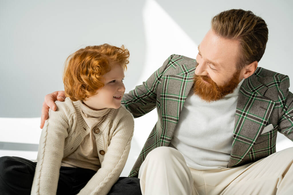 Uśmiechnięty brodaty mężczyzna przytulający rudego syna w swetrze na szarym tle z jasnym  - Zdjęcie, obraz