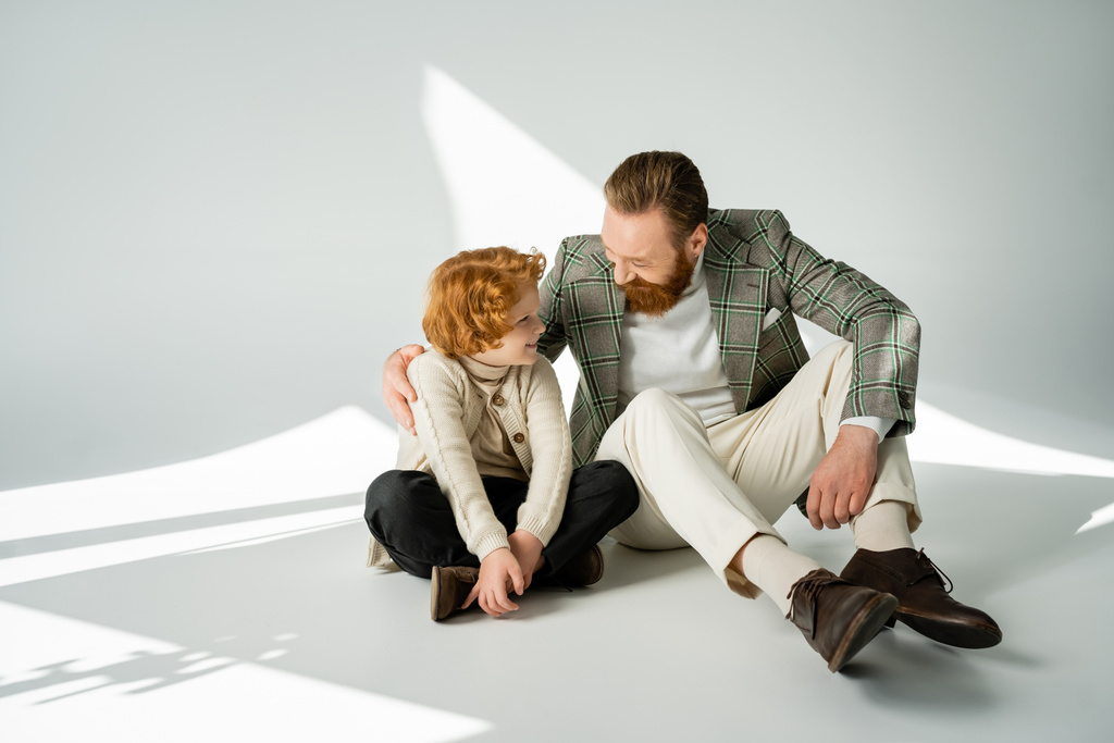 Positive man in jacket hugging red haired son while sitting on grey background with light  - Photo, Image
