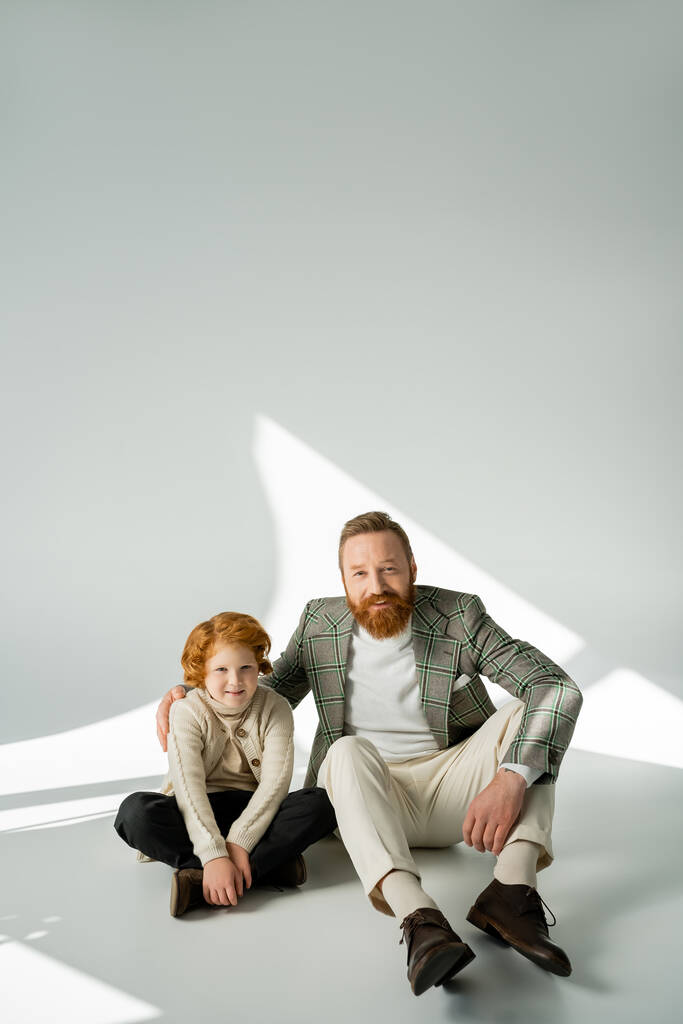 Padre e hijo de pelo rojo positivo mirando a la cámara mientras están sentados sobre un fondo gris con luz  - Foto, imagen