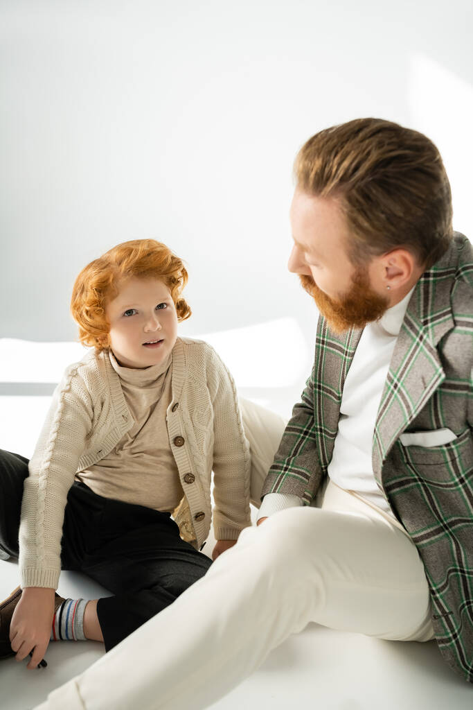 Red haired boy in jumper sitting near stylish dad on grey background with sunlight  - Photo, Image