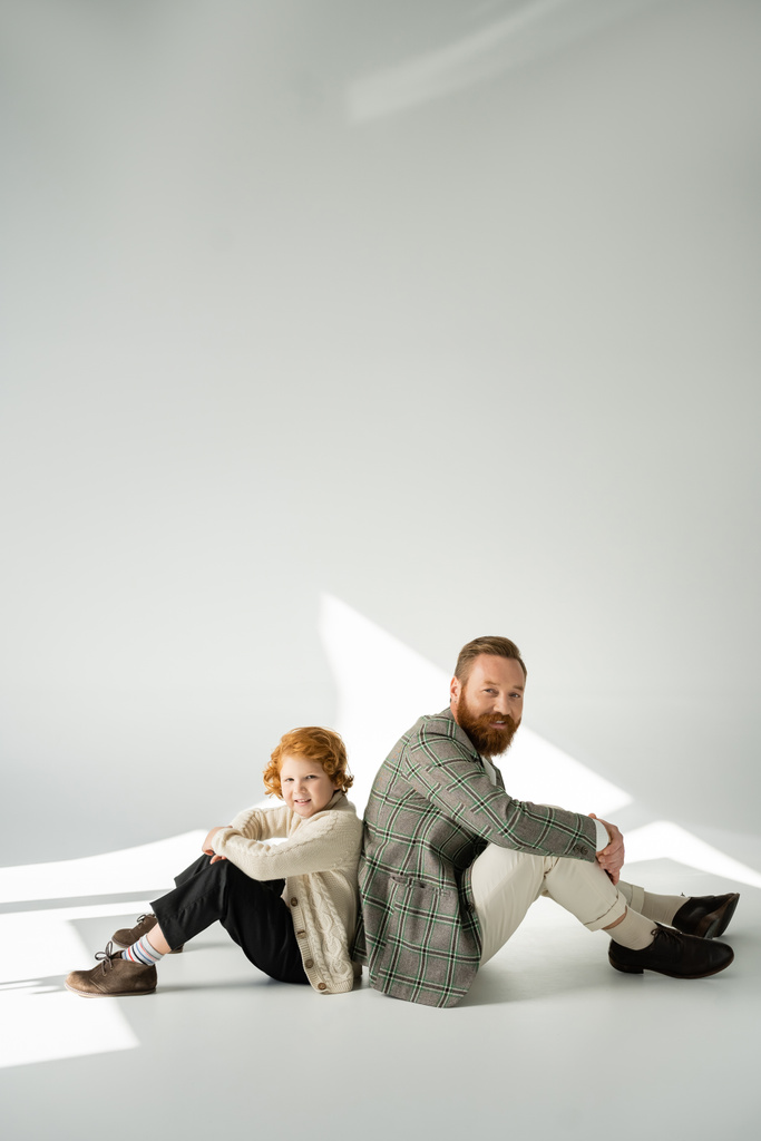 Stylish red haired kid and father sitting back to back on grey background with sunlight  - Photo, Image