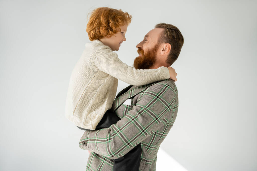 Side view of positive and bearded dad lifting son in knitted jumper on grey background - Photo, Image