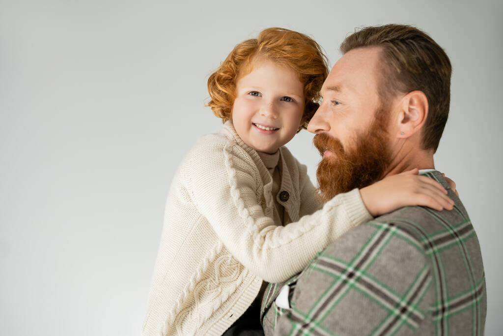 Red haired boy in knitted jumper hugging bearded father and looking at camera isolated on grey  - Photo, Image