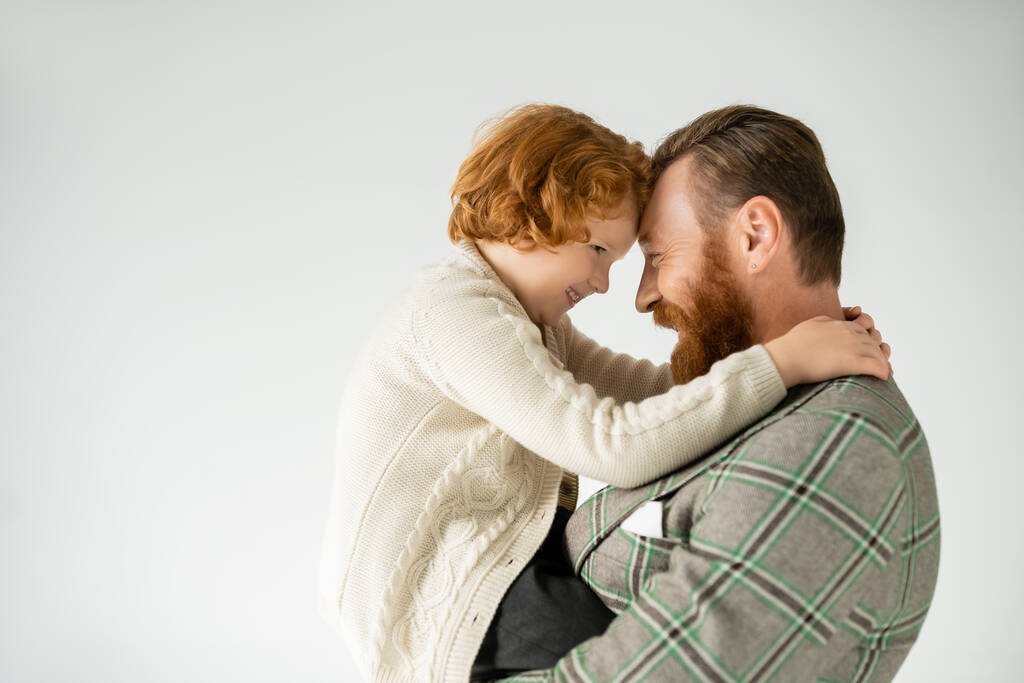 Πλευρική άποψη του χαμογελαστού κοκκινομάλλα παιδί αγκαλιάζει κομψό μπαμπά απομονωμένο σε γκρι  - Φωτογραφία, εικόνα