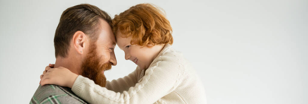 Seitenansicht eines positiven rothaarigen Kindes im gemütlichen Pullover, das den Vater isoliert auf grau umarmt, Banner  - Foto, Bild