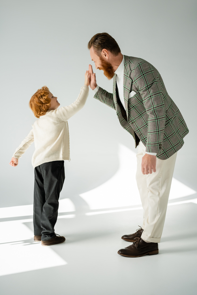 Vista lateral do homem positivo e elegante dando alta cinco para o filho de cabelos vermelhos no fundo cinza - Foto, Imagem