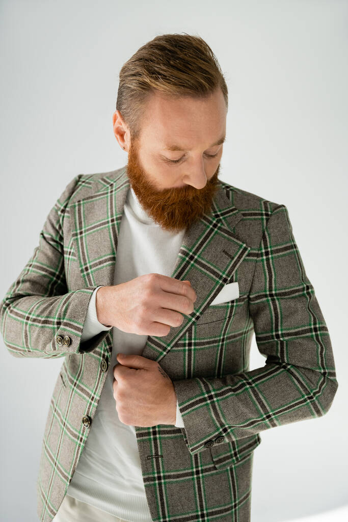 Elegante hombre barbudo mirando solapa de chaqueta sobre fondo gris - Foto, Imagen
