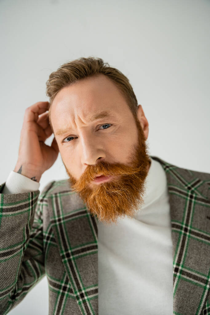 Портрет стильного и бородатого мужчины, трогающего волосы и смотрящего на камеру, изолированную на сером  - Фото, изображение