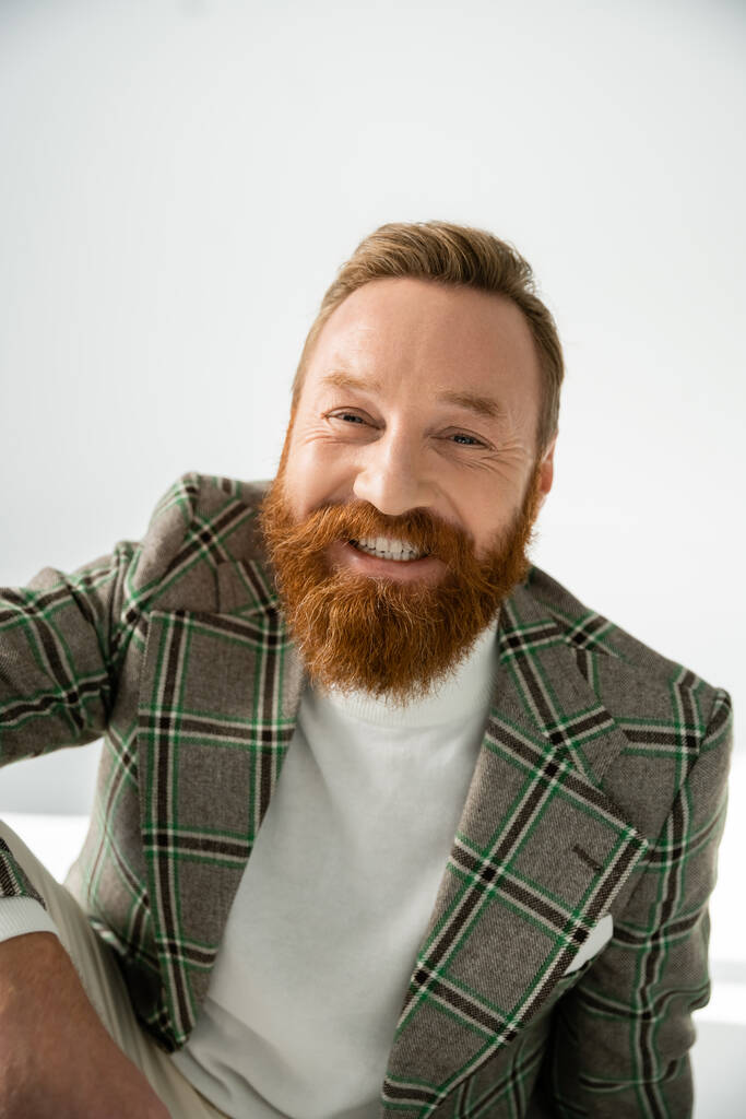 Позитивный бородатый мужчина в клетчатой куртке смотрит в камеру на сером фоне - Фото, изображение