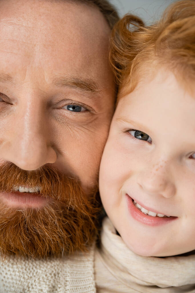 Vue recadrée d'un homme barbu souriant et d'un fils roux regardant une caméra isolée sur du gris  - Photo, image