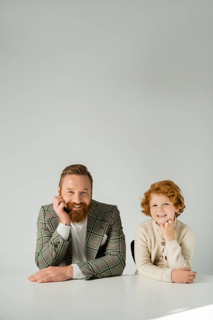 Smiling dad and red haired son looking at camera near table with sunlight isolated on grey  - Photo, Image