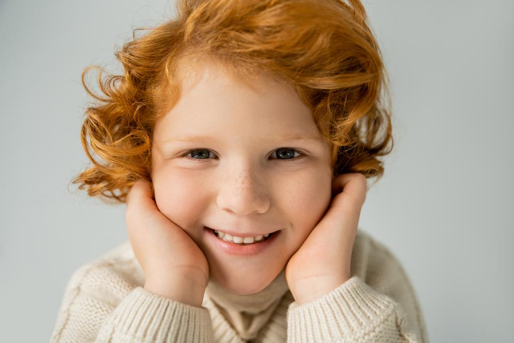 Cheerful red haired kid in beige jumper touching cheeks isolated on grey  - Photo, Image