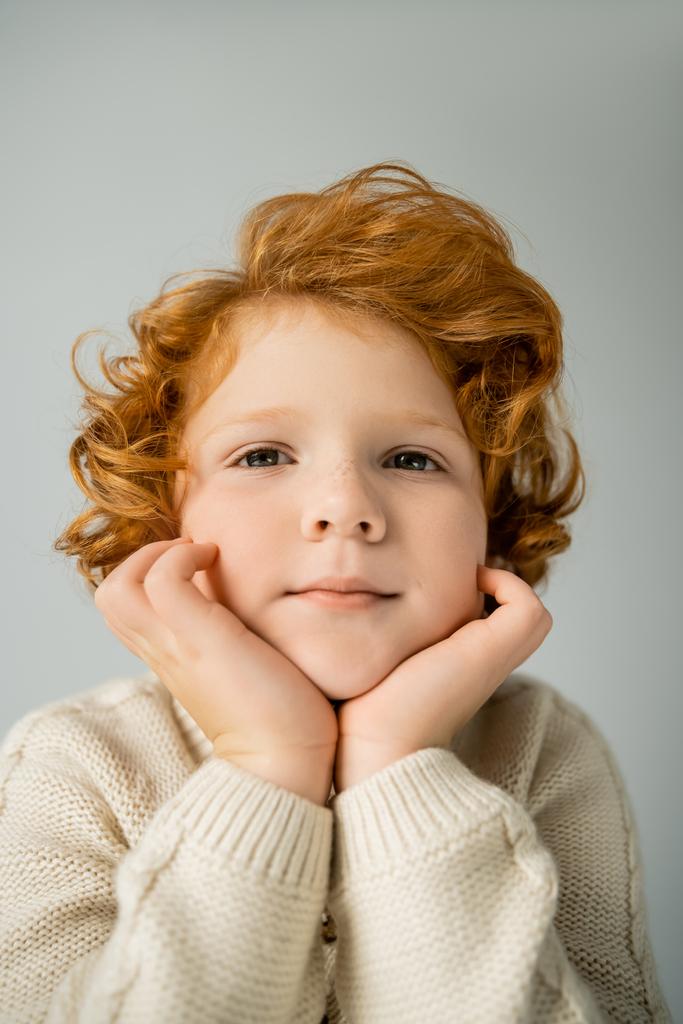 Rudowłosy chłopiec w ciepłym beżowym swetrze patrząc na aparat odizolowany na szarości  - Zdjęcie, obraz