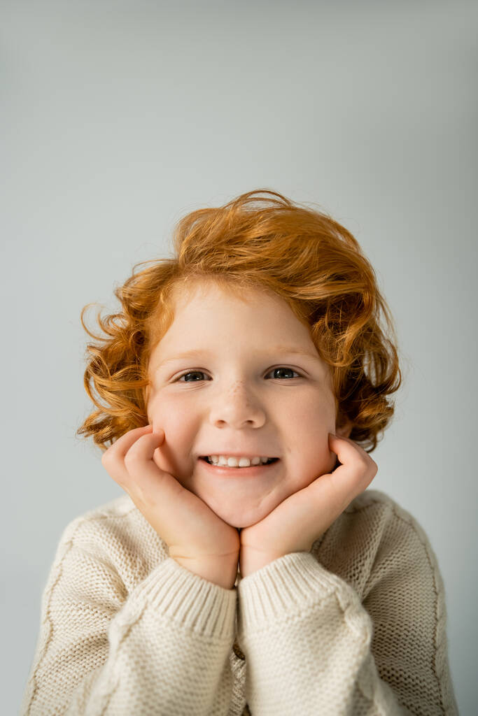 Red haired kid in warm knitted jumper smiling at camera isolated on grey  - Photo, Image