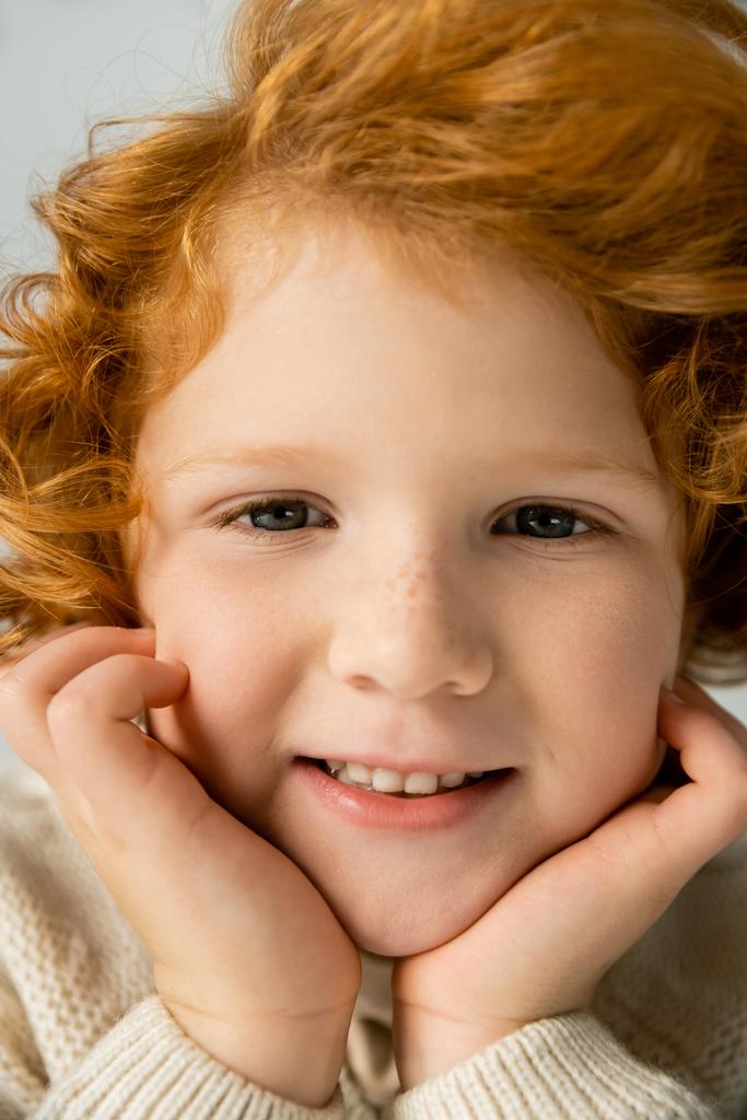 Nahaufnahme eines lächelnden rothaarigen Kindes im Strickpullover, das Wangen berührt, isoliert auf grau  - Foto, Bild