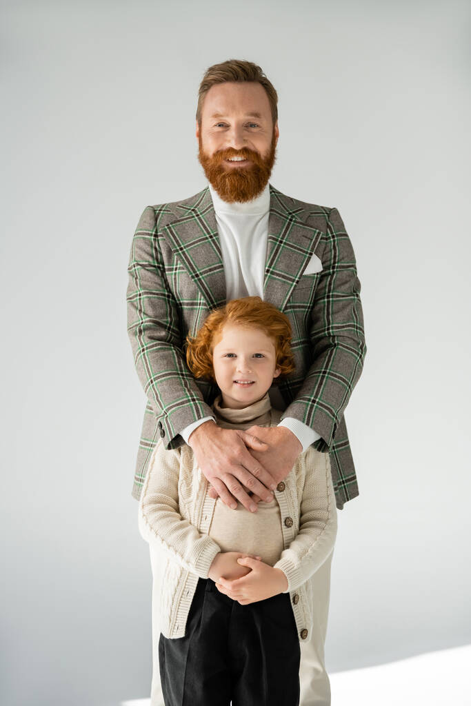 Ekose ceketli mutlu sakallı adam kızıl saçlı oğlanı kucaklıyor ve gri arka planda kameraya gülümsüyor. - Fotoğraf, Görsel