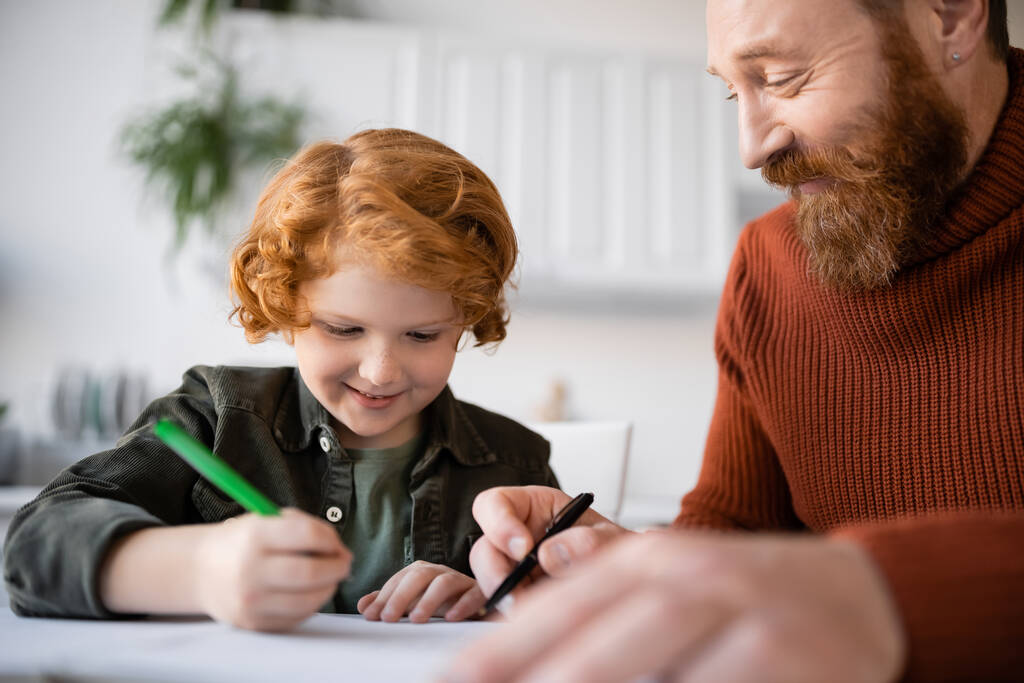 sorrindo criança ruiva escrevendo no caderno enquanto fazendo lição de casa perto do pai satisfeito - Foto, Imagem