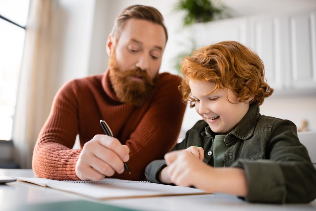 man met baard houden pen in de buurt notebook en glimlachende roodharige zoon doet huiswerk - Foto, afbeelding