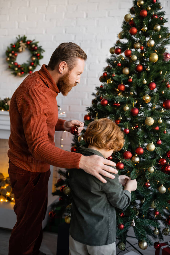 家でクリスマスツリーを飾るレッドヘッドの息子の近くに立つ髭の男 - 写真・画像