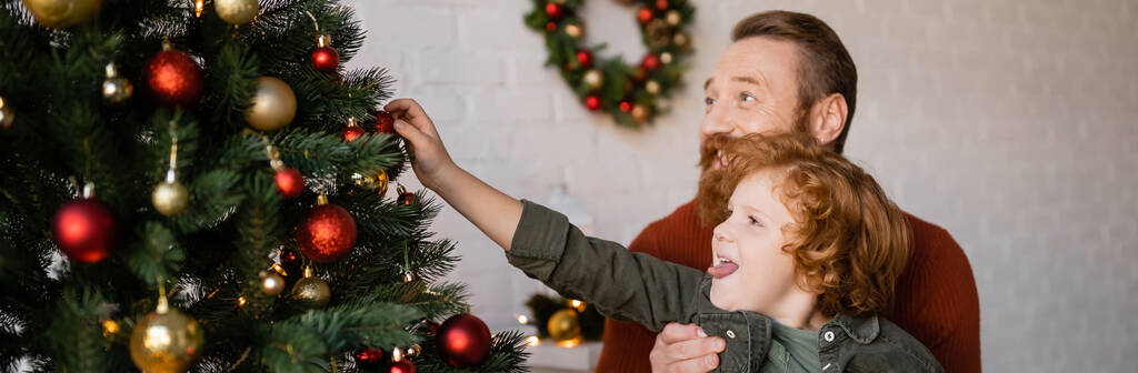 redhead boy sticking out tongue and decorating Christmas tree near bearded father, banner - Photo, Image
