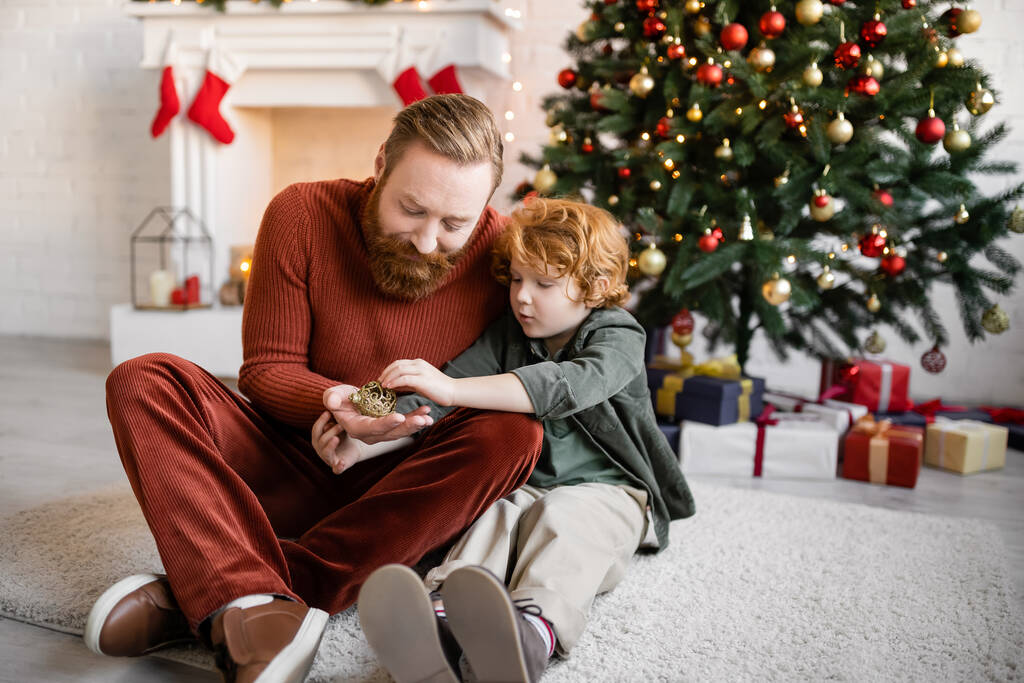 rousse père et enfant tenant boule de Noël sur le sol dans le salon avec décor festif - Photo, image