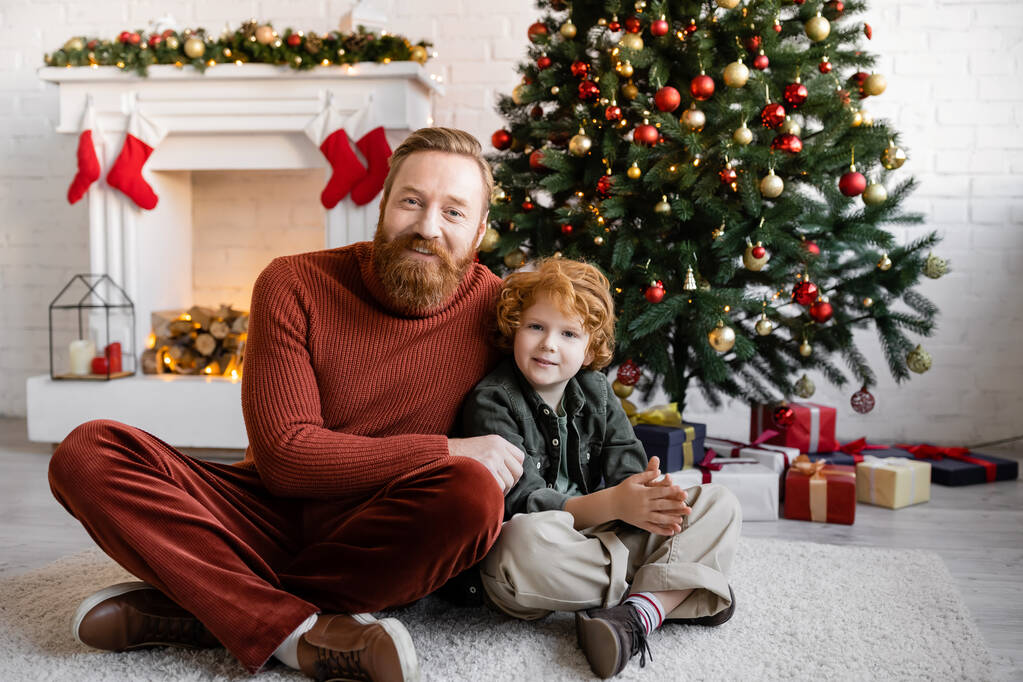 pelirroja padre e hijo sentado con las piernas cruzadas en el suelo en la sala de estar con árbol de Navidad y chimenea decorada - Foto, Imagen