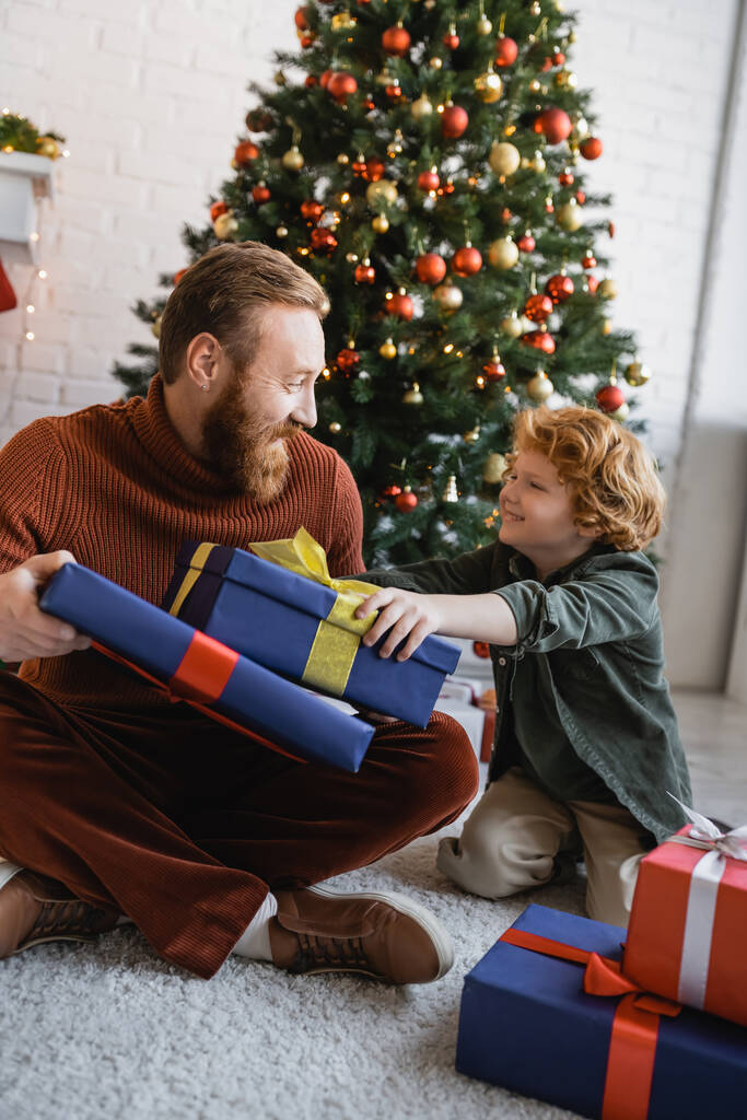 щасливий рудий батько і син дивляться один на одного біля подарункових коробок і ялинки вдома
 - Фото, зображення