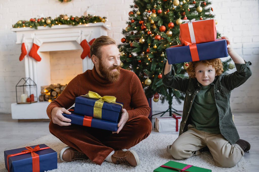 child holding gift boxes above head while having fun near bearded dad sitting on floor near Christmas tree - Photo, Image