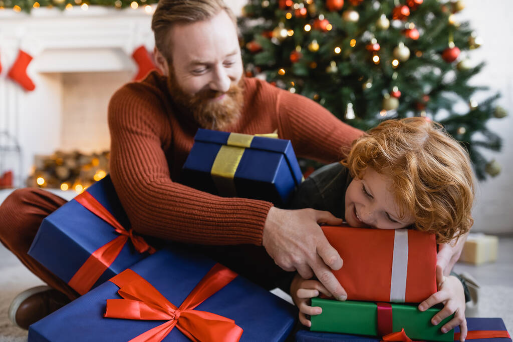陽気な髭を生やした男と赤毛の子供は家でクリスマスプレゼントボックスで楽しんでいます - 写真・画像